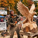 A chainsaw artist carving a wooden eagle sculpture at Hoos'Kow Chainsaw Carving 2026 outdoor festival in October
