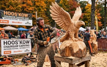 A chainsaw artist carving a wooden eagle sculpture at Hoos'Kow Chainsaw Carving 2026 outdoor festival in October